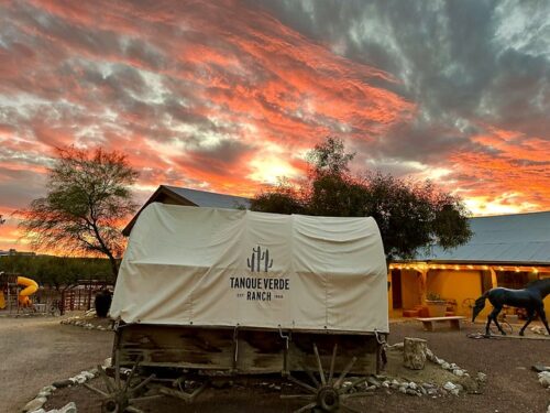 Canvas covered wagon on the grounds of Tanque Verde Ranch in Tucson, Arizona. The canvas wagon cover bears the logo Tanque Verde Ranch, established 1868, with a trio of 3 saguaro cactus on the beige fabric. An evening sunset lights up the sky with clouds of pink and grey. Two scraggly trees flank the wagon with the stables building partially blocked, behind the antique chuck wagon.